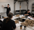 Adults debating around a workshop table covered with papers and untouched coils of rope while an empty suspension frame stands behind them.