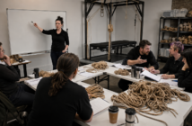 Adults debating around a workshop table covered with papers and untouched coils of rope while an empty suspension frame stands behind them.